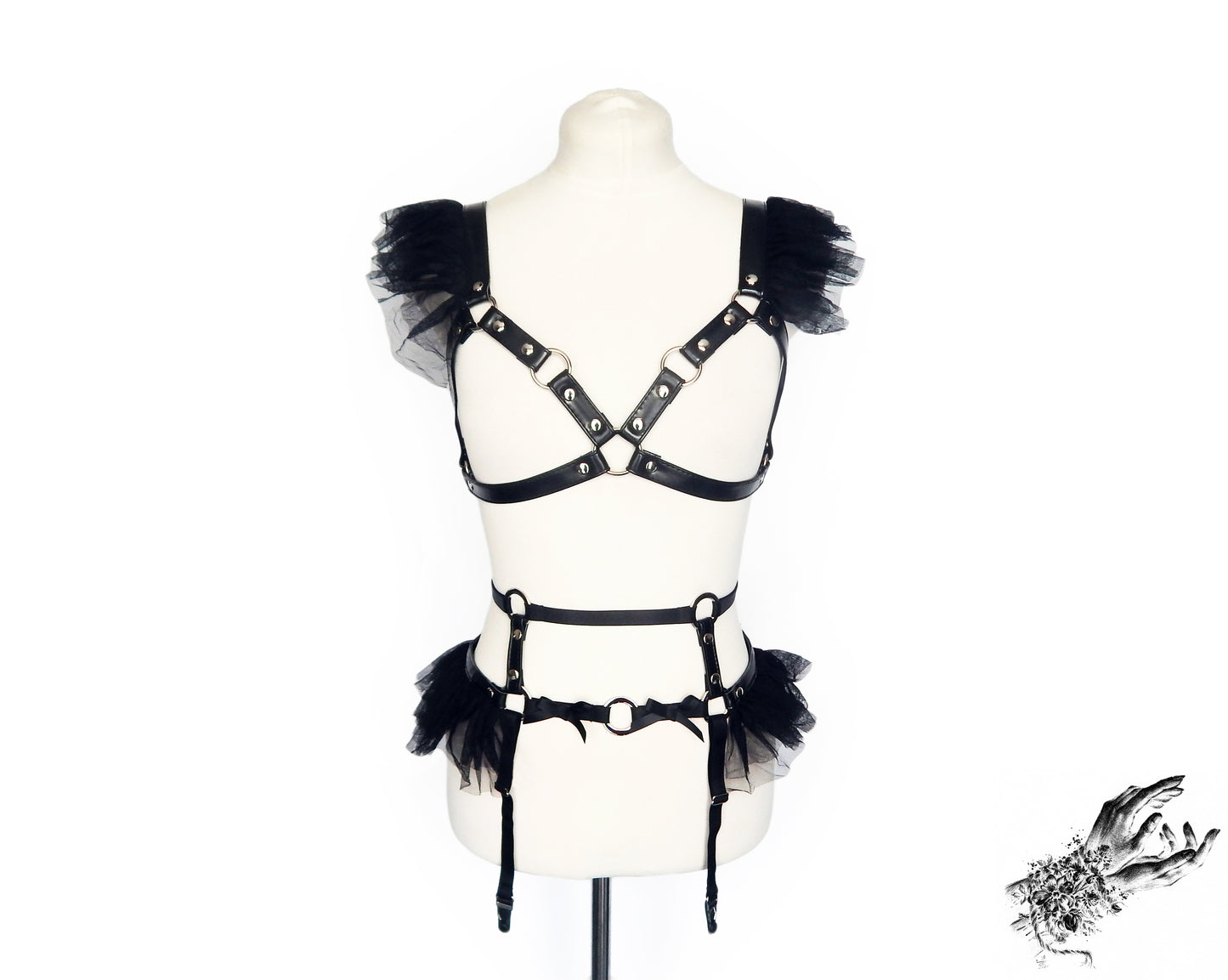 Front view of a black vegan leather harness set featuring a separate harness bra and garter belt with silver O ring hardware, ruffled tulle and bow appliques, displayed on a mannequin against a white background.