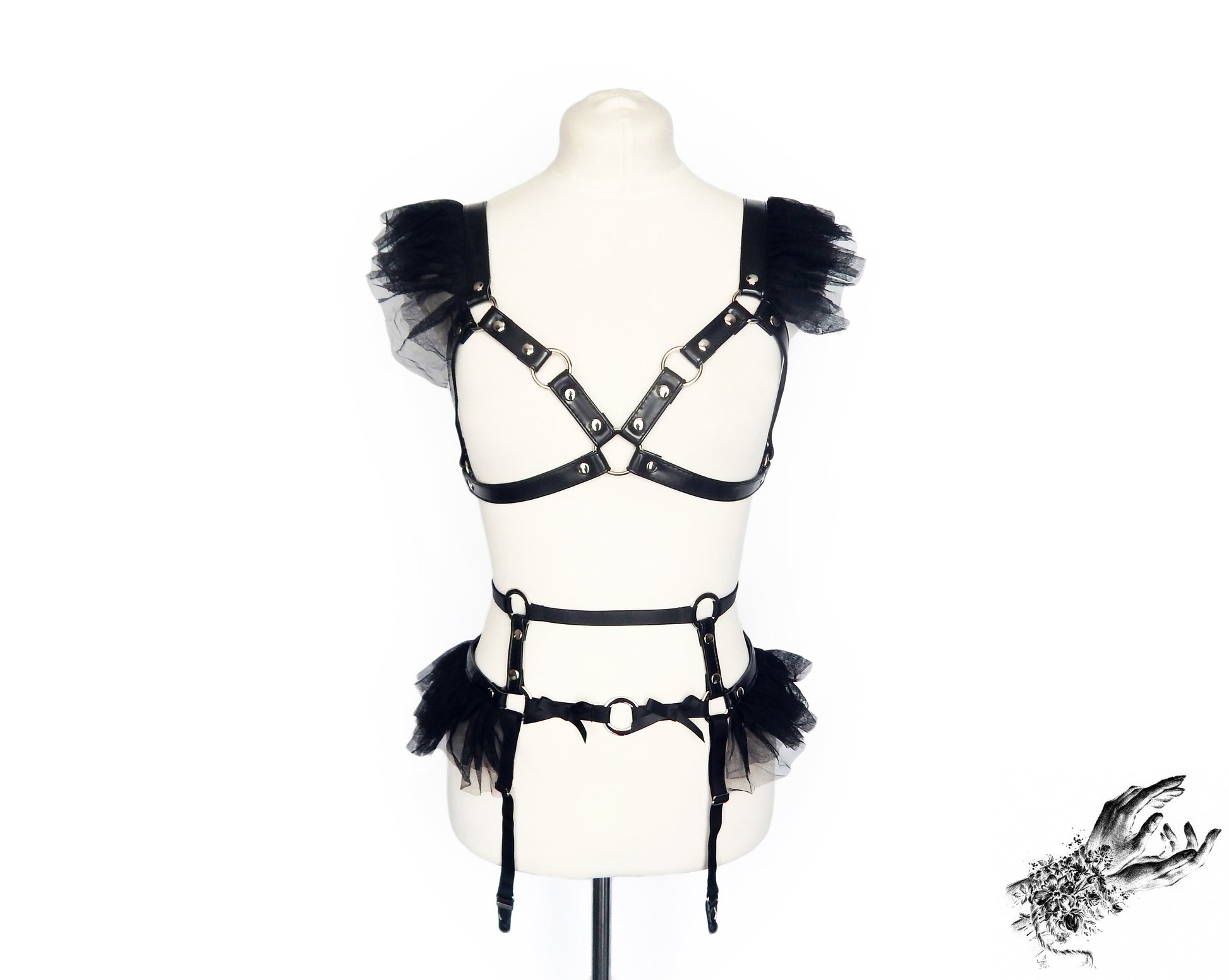 Front view of a black vegan leather harness set featuring a separate harness bra and garter belt with silver O ring hardware, ruffled tulle and bow appliques, displayed on a mannequin against a white background.