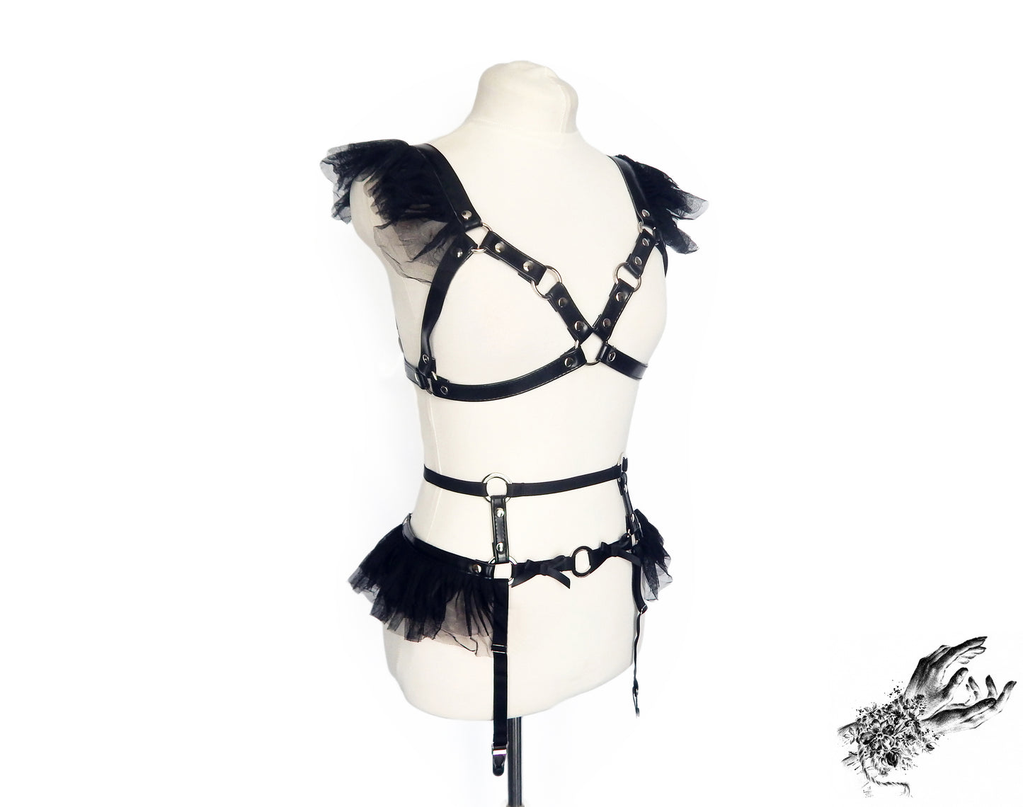 Black vegan leather harness set featuring a separate harness bra and garter belt with silver O ring hardware, ruffled tulle and bow appliques, displayed on a mannequin against a white background.