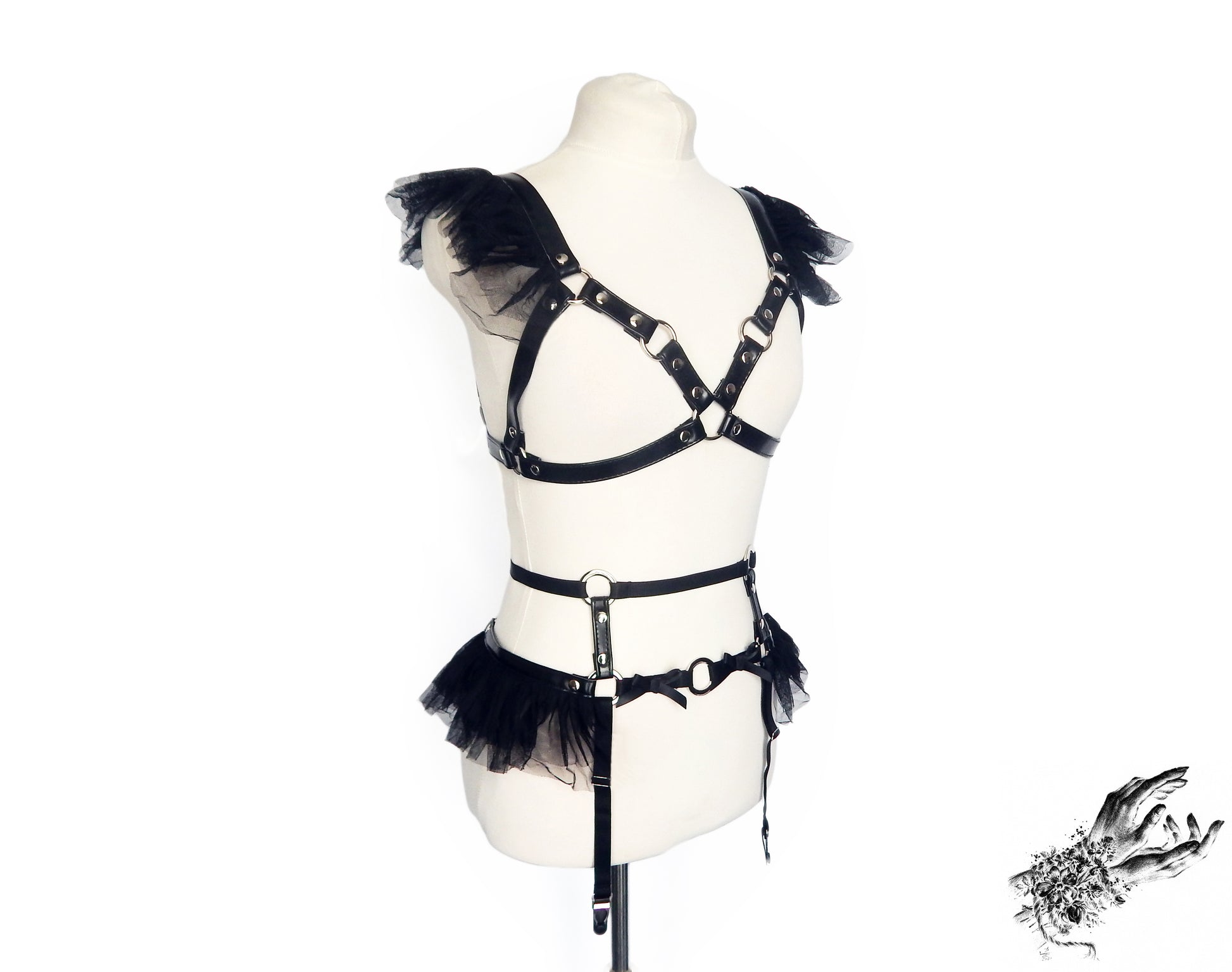 Black vegan leather harness set featuring a separate harness bra and garter belt with silver O ring hardware, ruffled tulle and bow appliques, displayed on a mannequin against a white background.