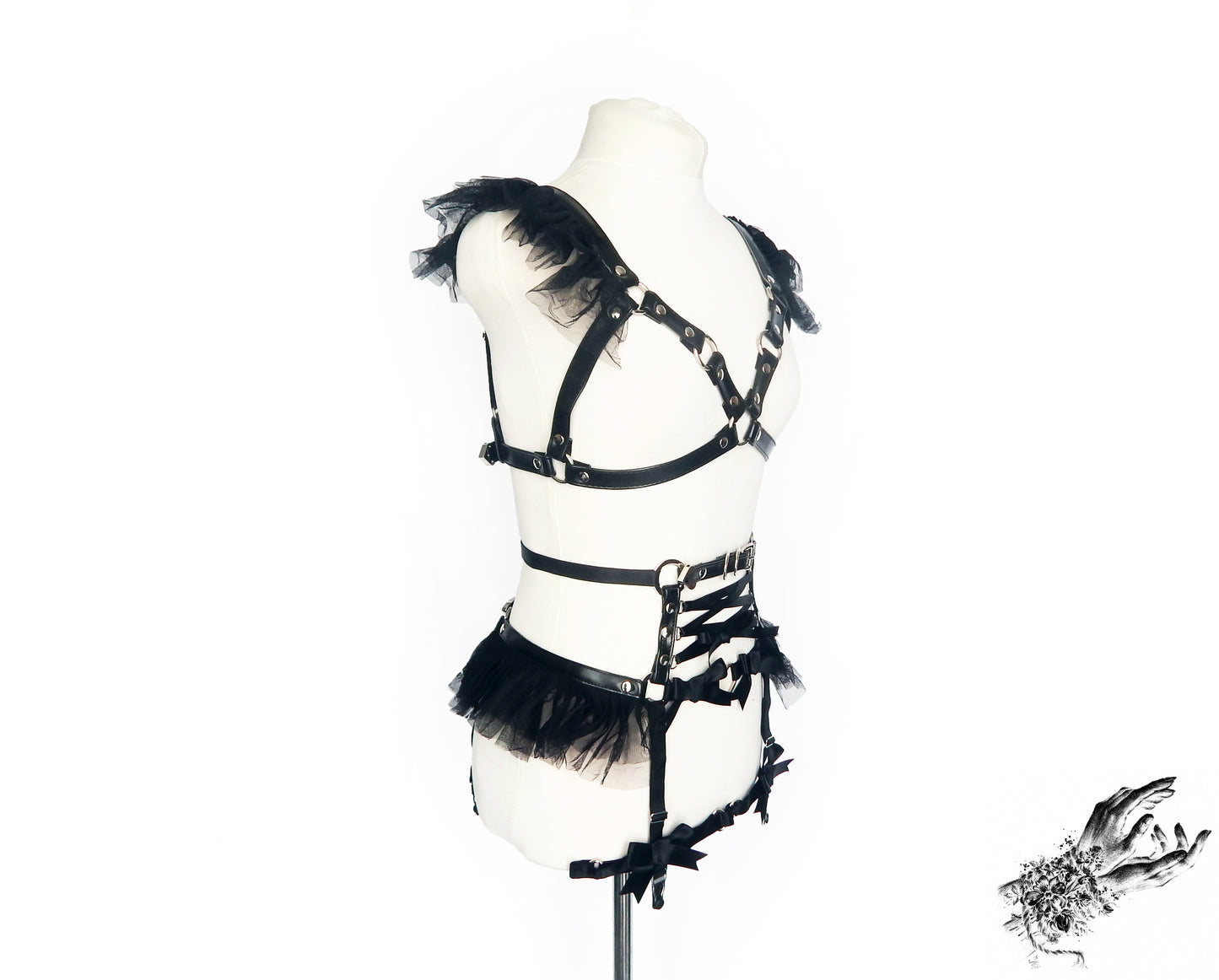 Side view of a black vegan leather harness set featuring a separate harness bra and garter belt with ruffled tulle accents, corset lacing and silver heart ring hardware, displayed on a mannequin against a white background.