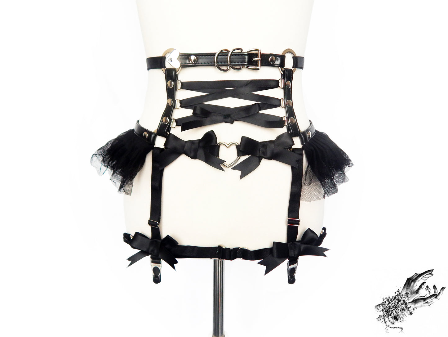 Black faux leather garter belt featuring central satin ribbon corset lacing, silver heart ring hardware, ruffled tulle and bow appliques, displayed on a mannequin against a white background.