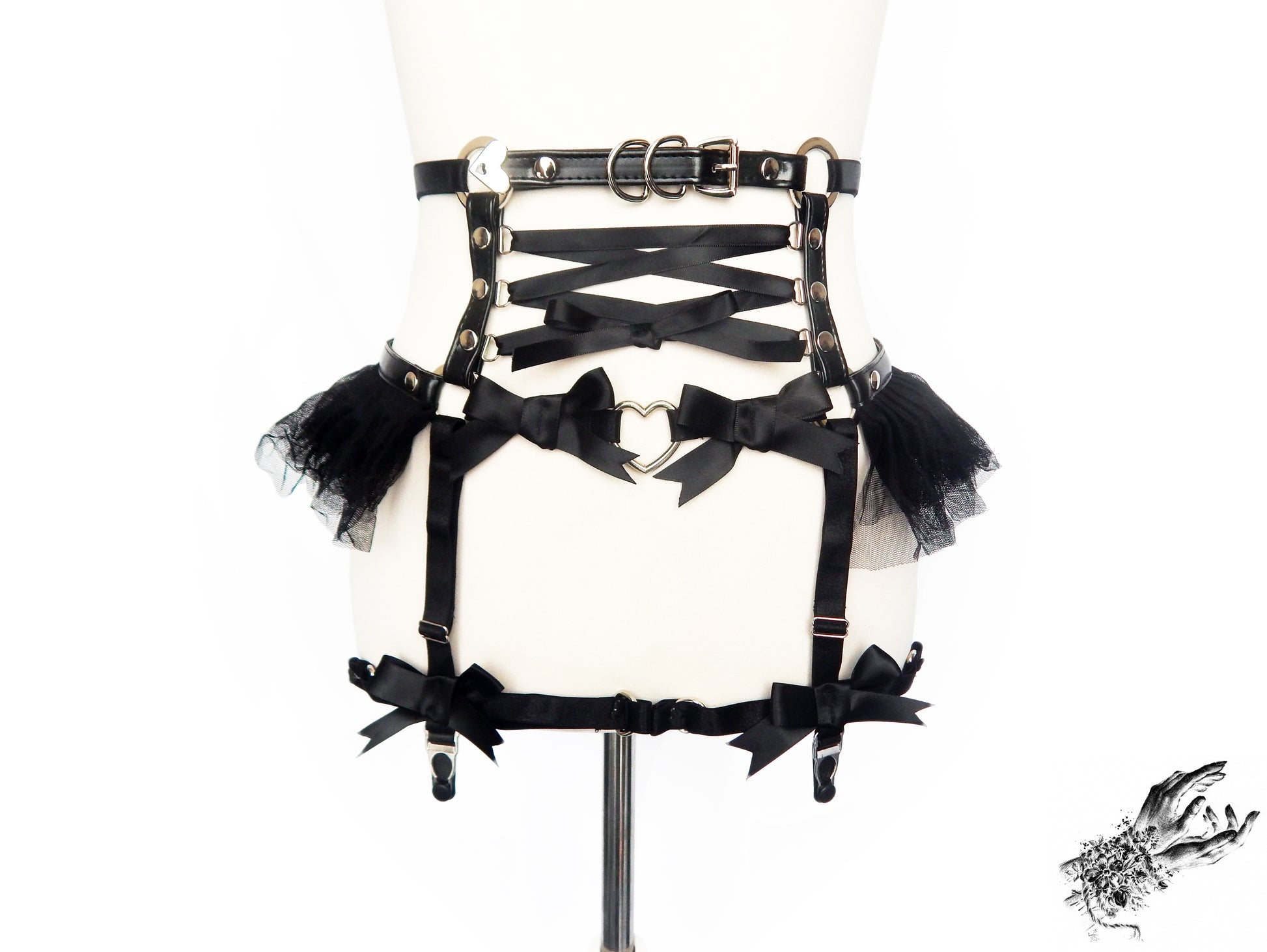 Black faux leather garter belt featuring central satin ribbon corset lacing, silver heart ring hardware, ruffled tulle and bow appliques, displayed on a mannequin against a white background.