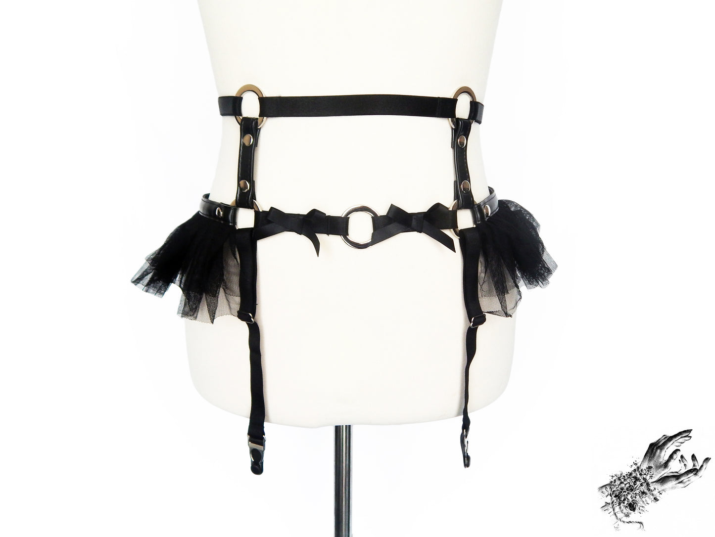 Black vegan leather garter belt featuring silver O ring hardware, ruffled tulle at the hips and bow appliques, displayed on a mannequin against a white background.