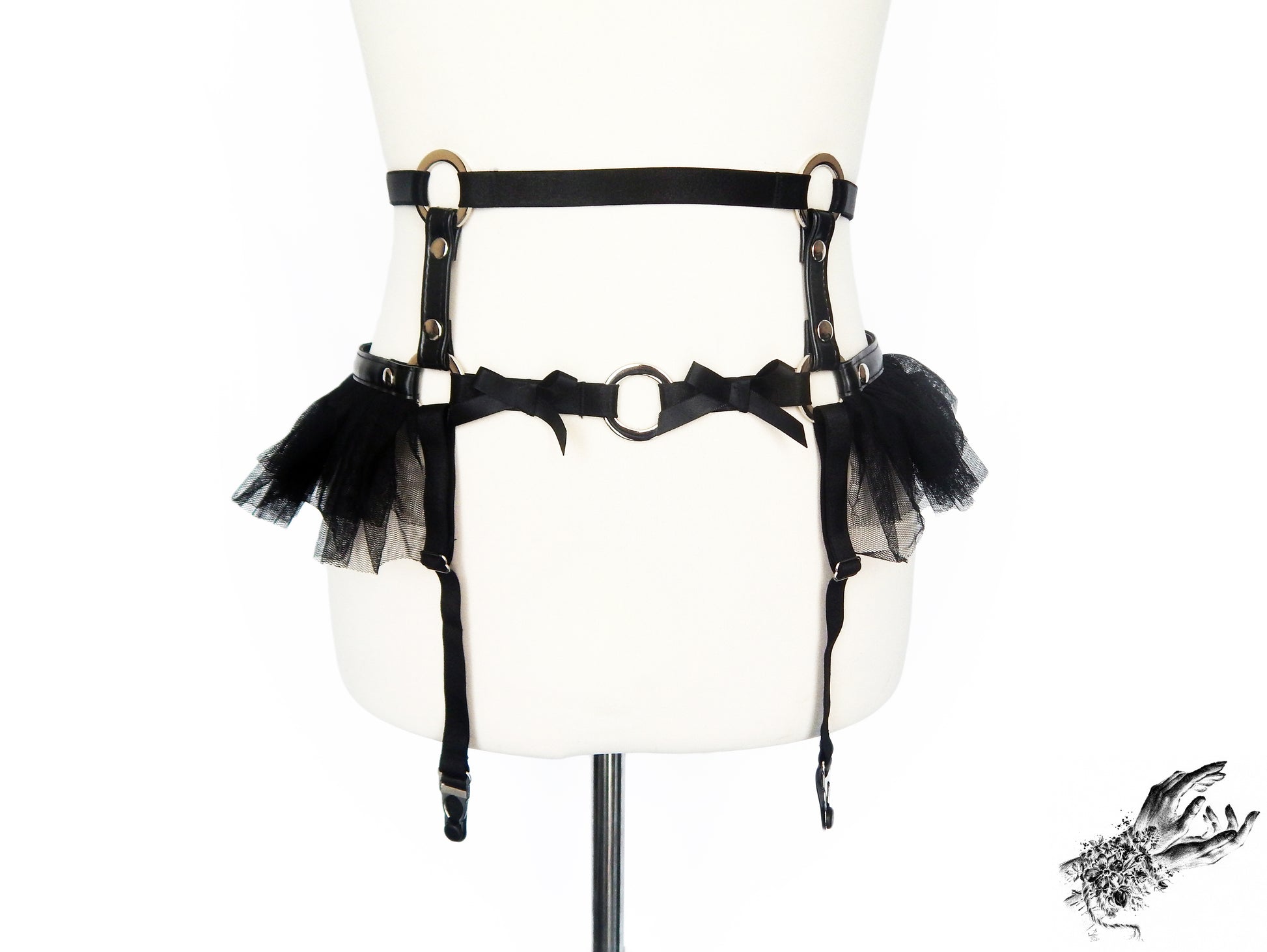 Black vegan leather garter belt featuring silver O ring hardware, ruffled tulle at the hips and bow appliques, displayed on a mannequin against a white background.