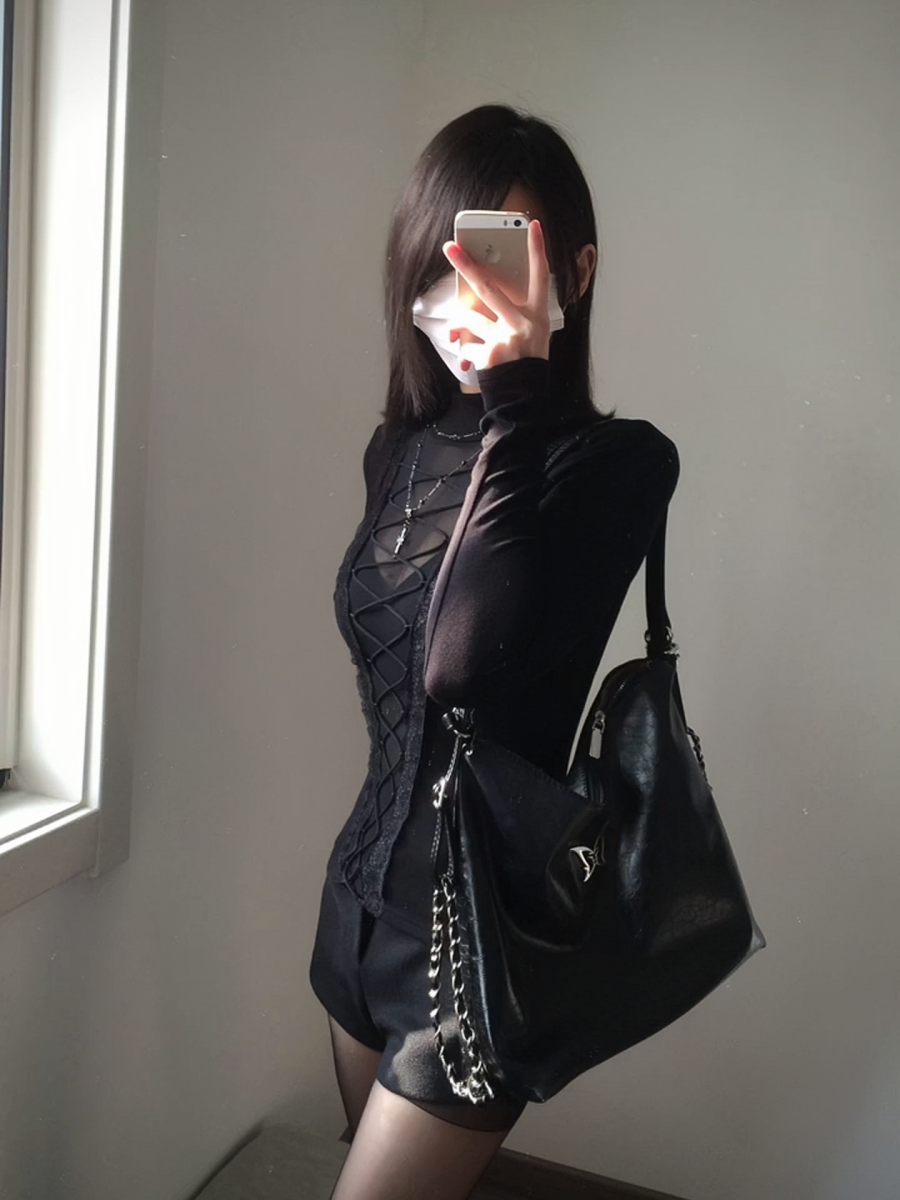 Modelled photo of a black lace-up top with turtleneck collar, sheer central bodice panel, long sleeves and elastic corset lacing. Styled with a silver cross necklace, black handbag and black shorts.
