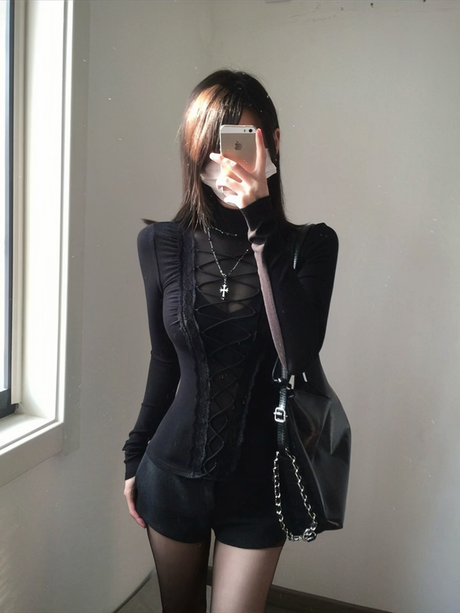 Modelled photo of a black lace-up top with turtleneck collar, sheer central bodice panel, long sleeves and elastic corset lacing. Styled with a silver cross necklace, black handbag and black shorts.