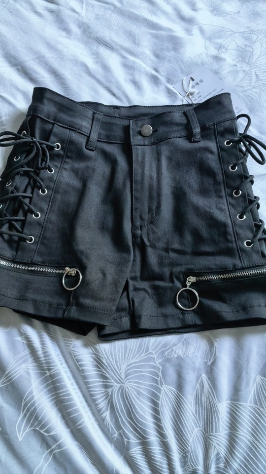 Black denim shorts with side lace-up details, metal O-ring hardware, and zippered cuffs, laid flat on a light gray patterned sheet.