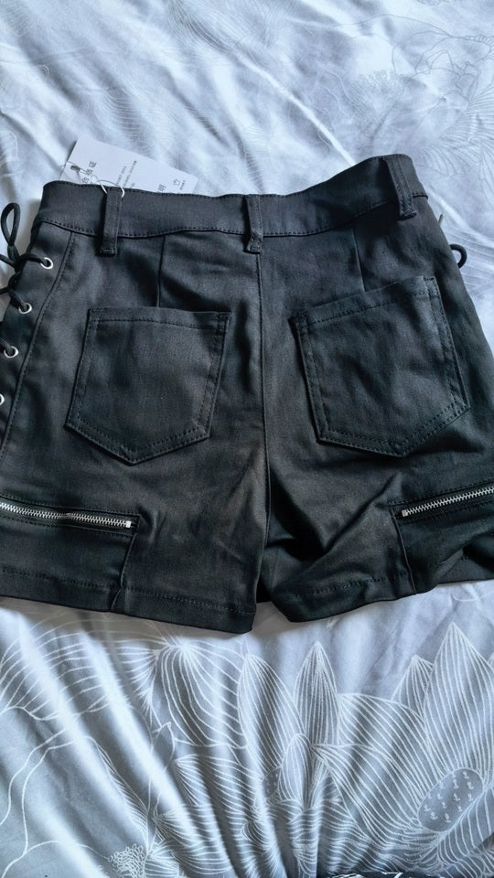 Black denim shorts with side lace-up detailing, rear patch pockets, and zippered cuffs, laid flat on a light gray patterned bedsheet.