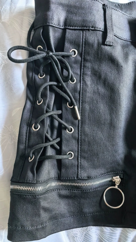A close-up of black denim shorts featuring a lace-up side panel, silver metal eyelets, and zippered cuff with an O ring pull, laid flat on a white textured fabric background.