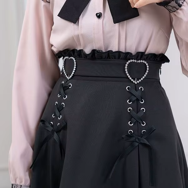 Model photo of a black high waisted skirt featuring a ruffled waist with side zip fastening, diamante heart shaped buckle belt, satin ribbon corset lacing and pleated a-line design, styled with a pink and black blouse for a Jirai Kei fashion aesthetic.