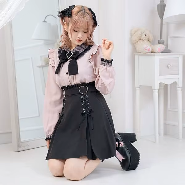Model photo of a black high waisted skirt featuring a ruffled waist, diamante heart shaped buckle belt, satin ribbon corset lacing and pleated a-line design, styled with a pink and black blouse and chunky black Mary Jane shoes for a Jirai Kei fashion aesthetic.