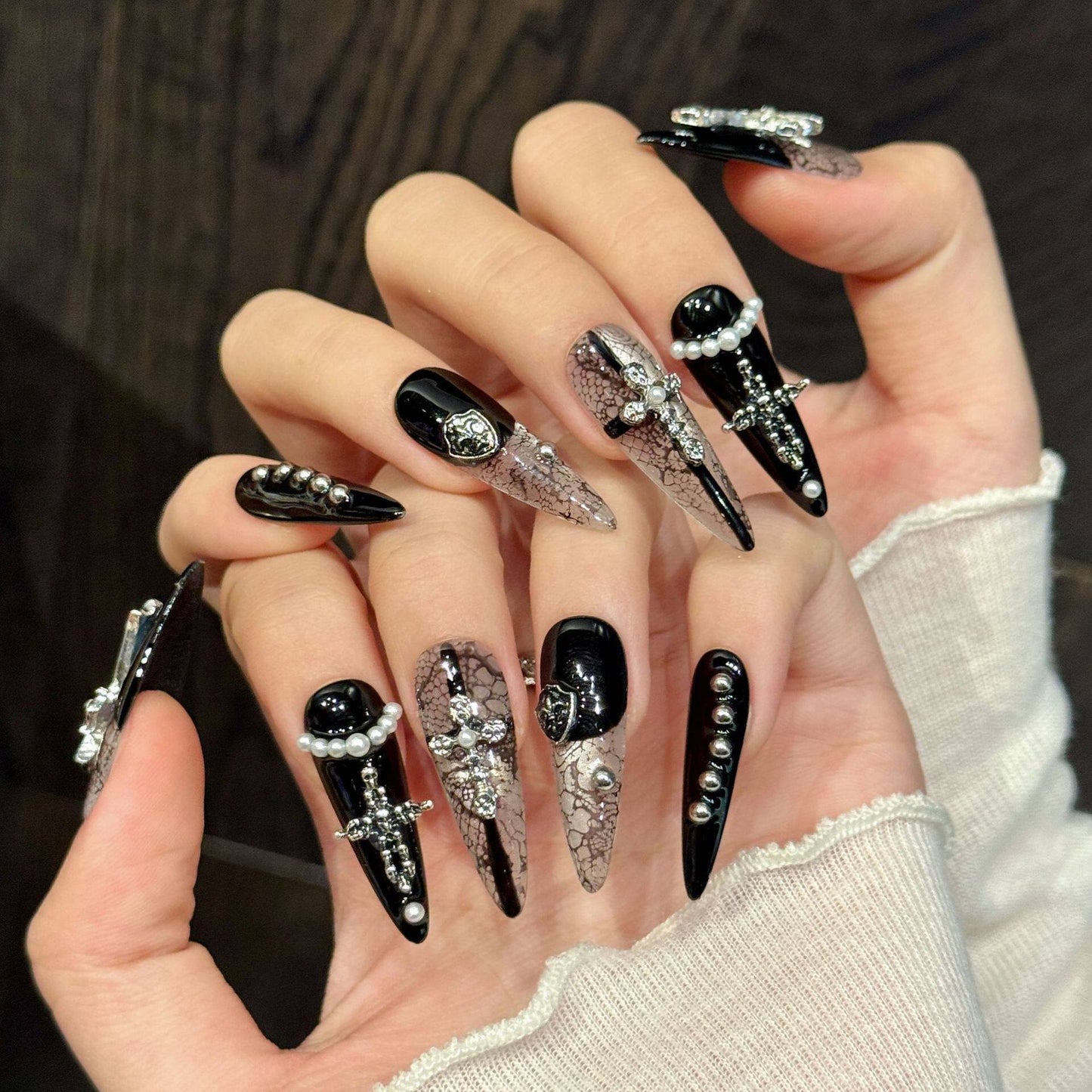 Black and white gothic snakeskin false nails in long stiletto length, featuring silver metal crosses and shields, white pearl beads and miniature studs, modelled on a hand.