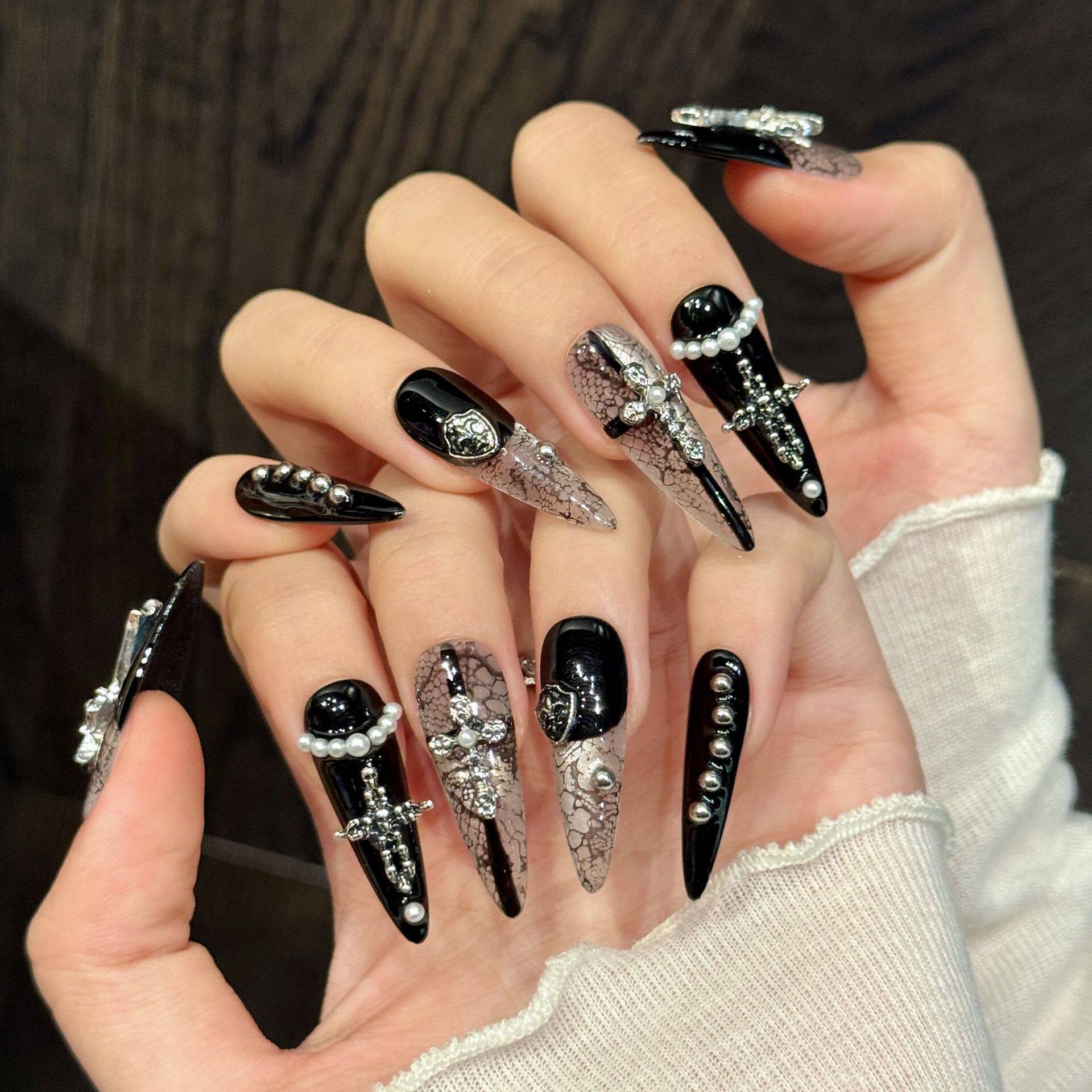 Black and white gothic snakeskin false nails in long stiletto length, featuring silver metal crosses and shields, white pearl beads and miniature studs, modelled on a hand.