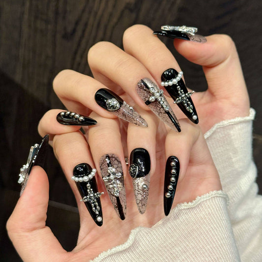 Black and white gothic snakeskin false nails in long stiletto length, featuring silver metal crosses and shields, white pearl beads and miniature studs, modelled on a hand.