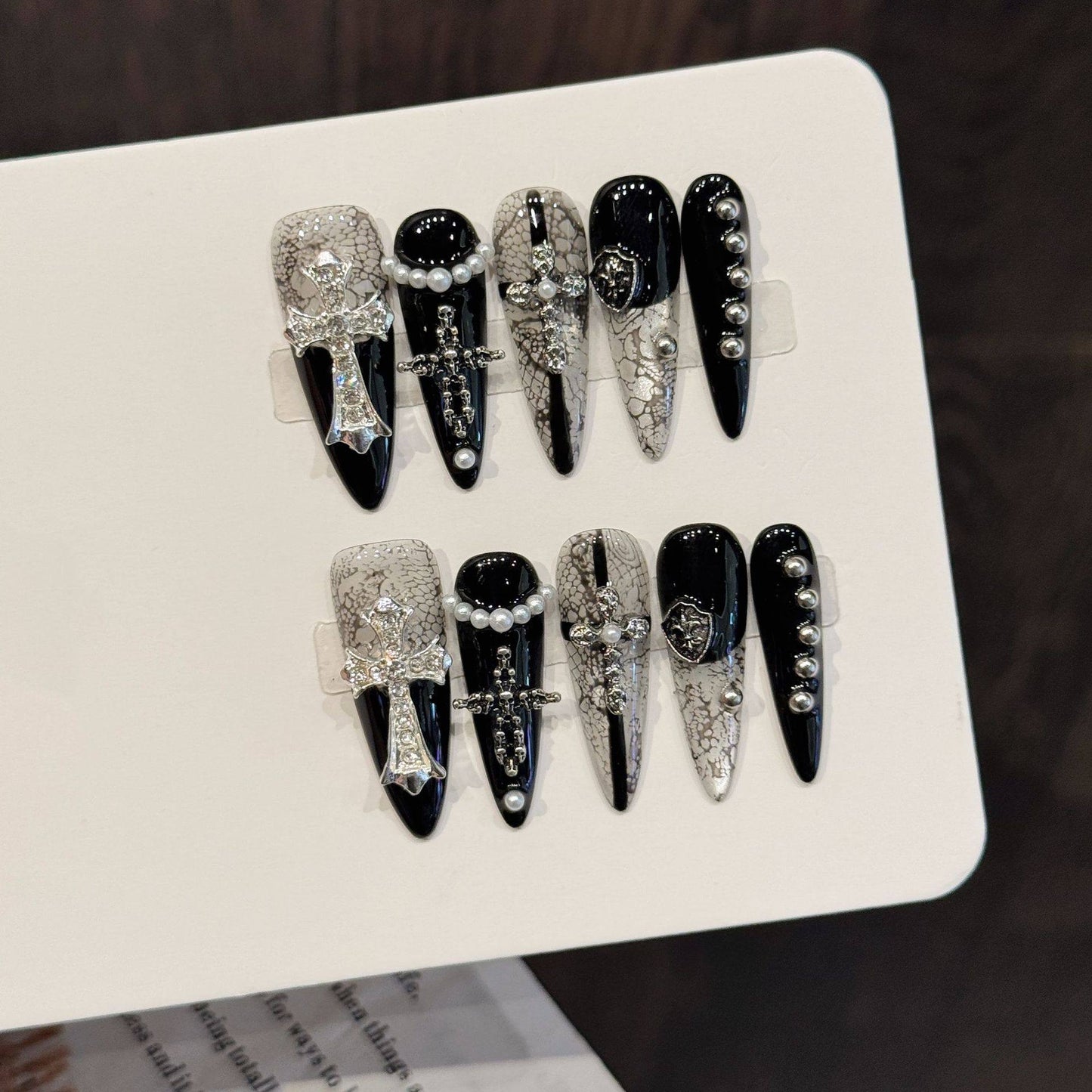 Black and white long stiletto false nails featuring a gothic snakeskin pattern, silver metal crosses and shields, white pearl beads and miniature studs, photographed on a white display card.