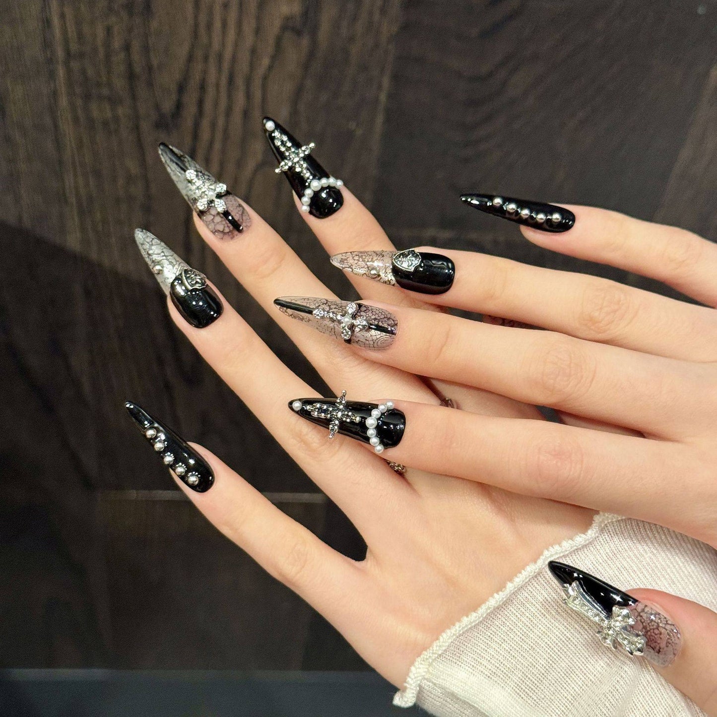 Black and white gothic snakeskin false nails in long stiletto length, featuring silver metal crosses and shields, white pearl beads and miniature studs, modelled on a hand.