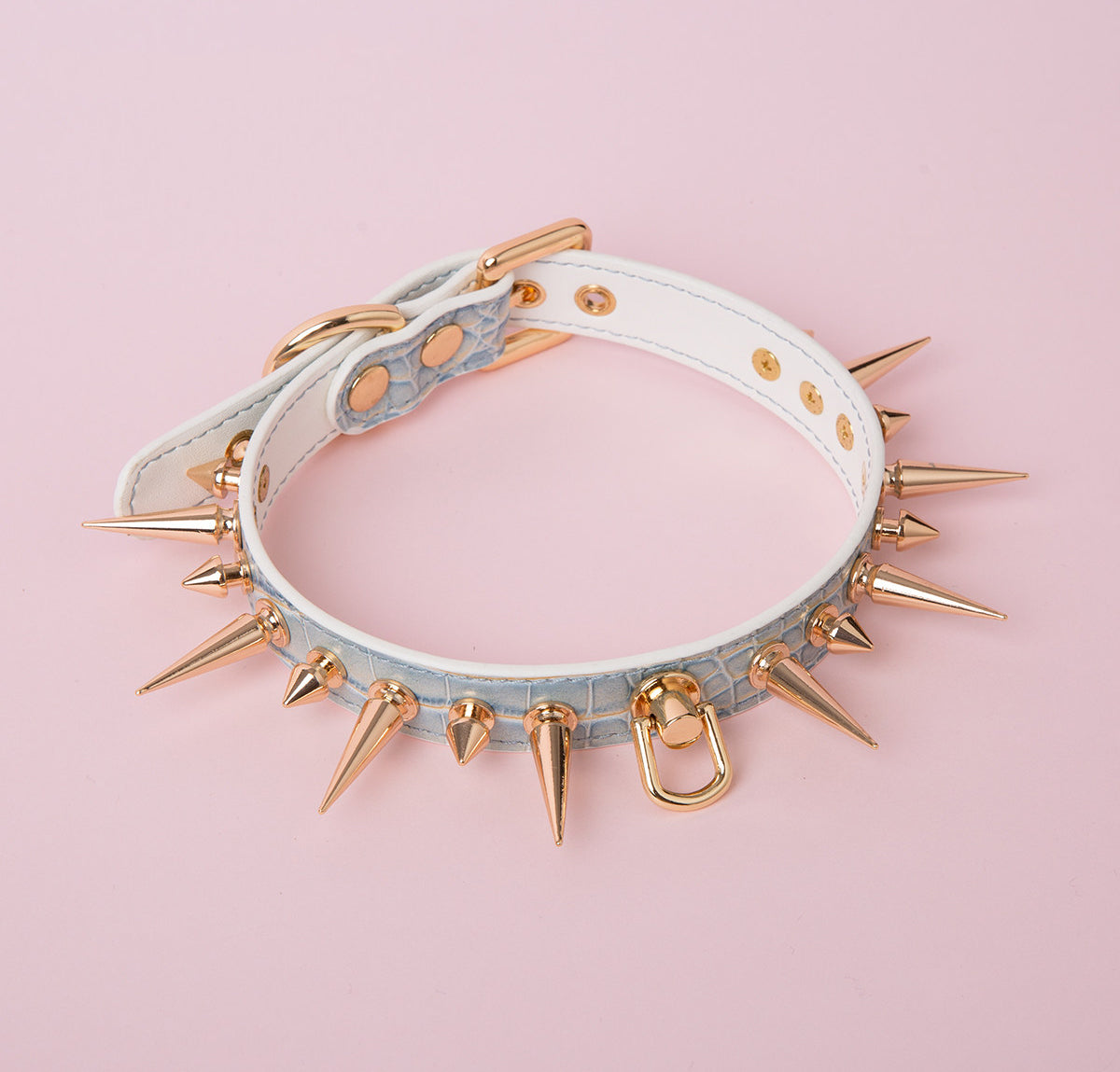 Blue iridescent faux crocodile effect leather choker featuring gold tone alternating length spike studs and central D ring attachment. Displayed on a pink background for a Harajuku fashion aesthetic.