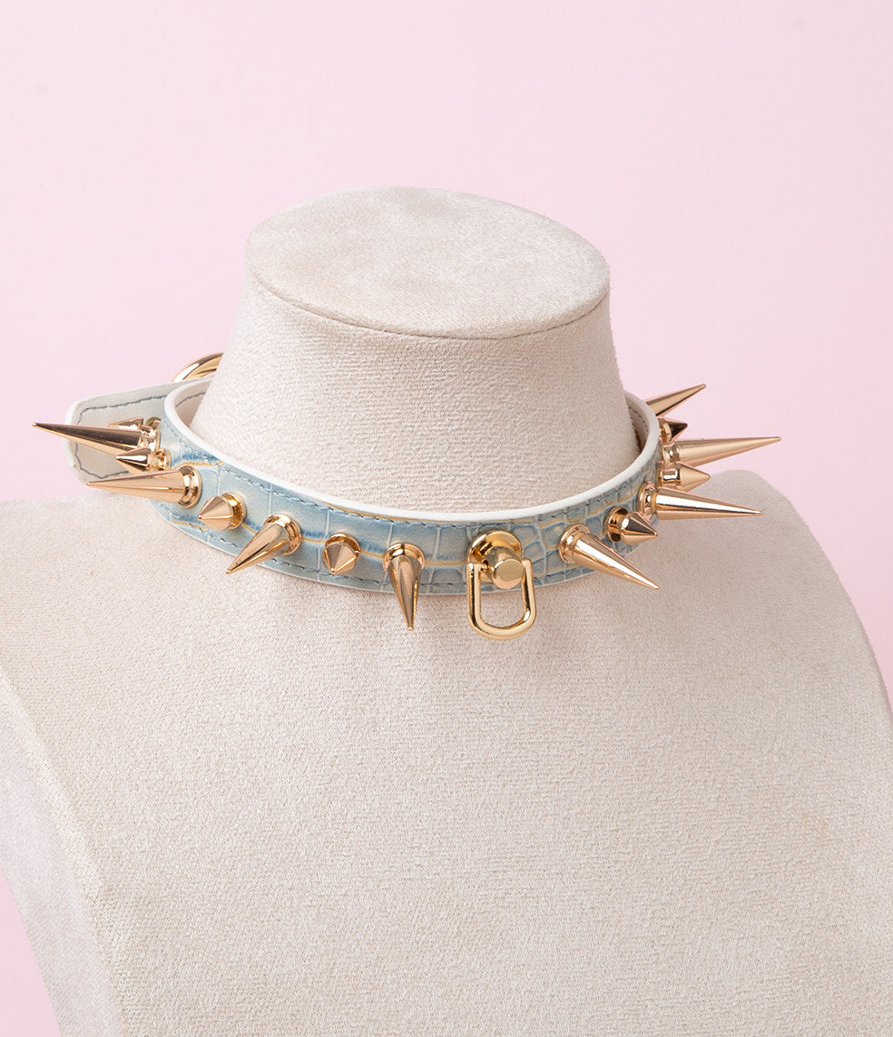 Blue iridescent faux crocodile effect leather choker featuring gold tone alternating length spike studs and central D ring attachment. Displayed on a white mannequin bust against a pink background for a kawaii goth aesthetic.