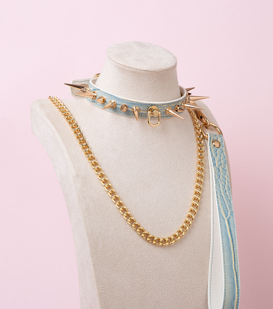 Blue faux crocodile effect leather choker and leash set featuring gold tone alternating length spike studs and central D ring attachment, with detachable chain leash and leather hand strap. Displayed on a white mannequin bust against a pink background for a pastel goth aesthetic.