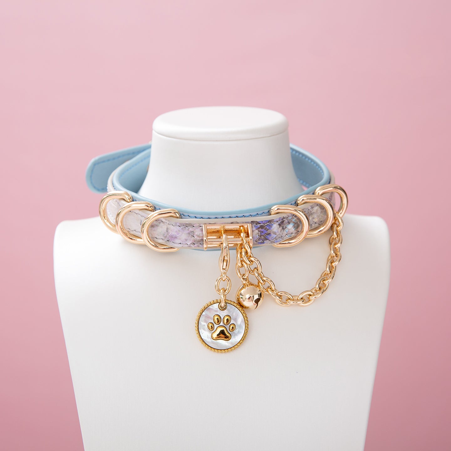 Baby blue leather choker featuring a gold and white paw print tag, gold tone D rings, chain and bell. Displayed on a white mannequin bust against a pink background for a Harajuku fashion aesthetic.
