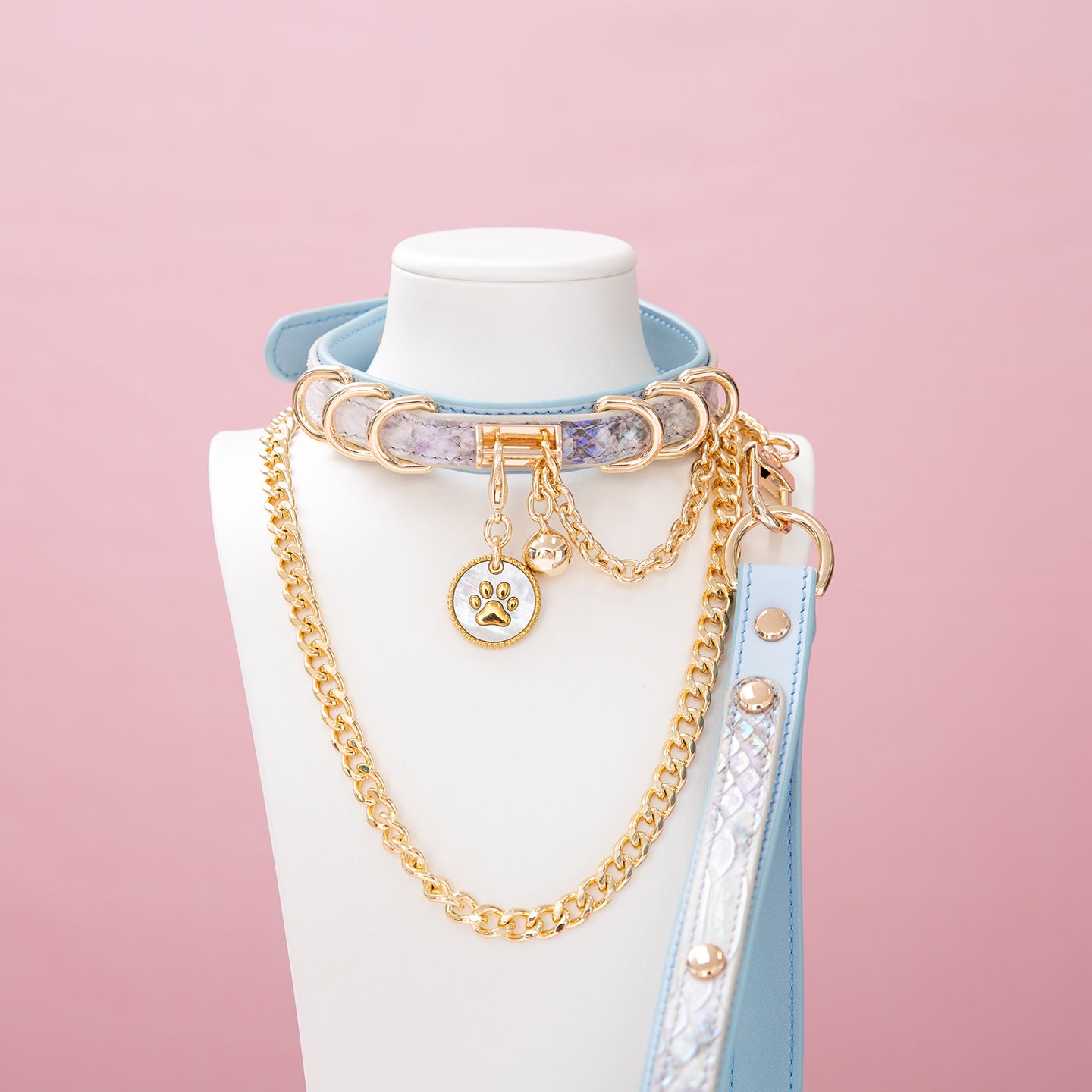 Baby blue leather choker and leash set featuring a white and gold paw print tag, D rings, chain and bell with detachable chain leash and leather hand strap. Displayed on a white mannequin bust against a pink background for a pastel goth aesthetic.