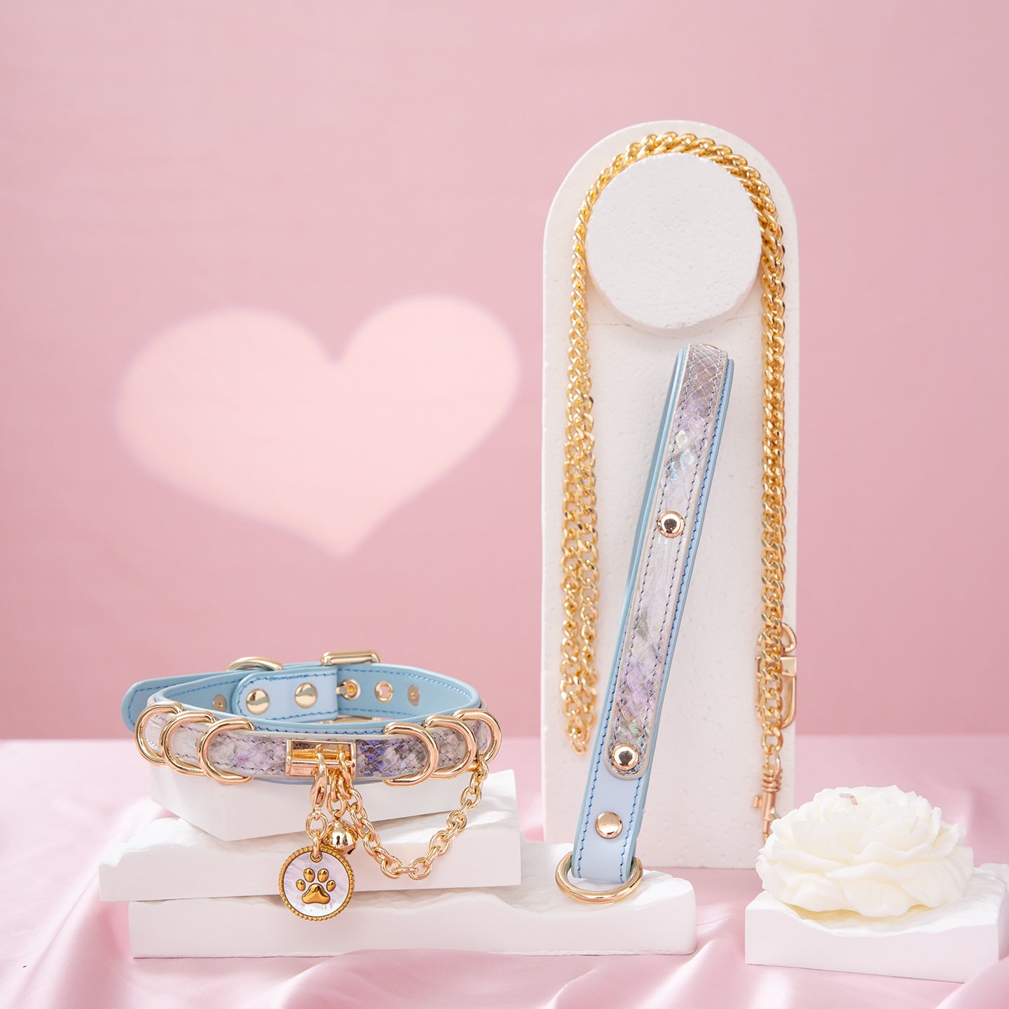 Baby blue leather choker and leash set featuring a white and gold paw print tag, D rings, chain and bell with detachable chain leash and leather hand strap. Displayed on a white display stand against a pink background for a pastel goth aesthetic.