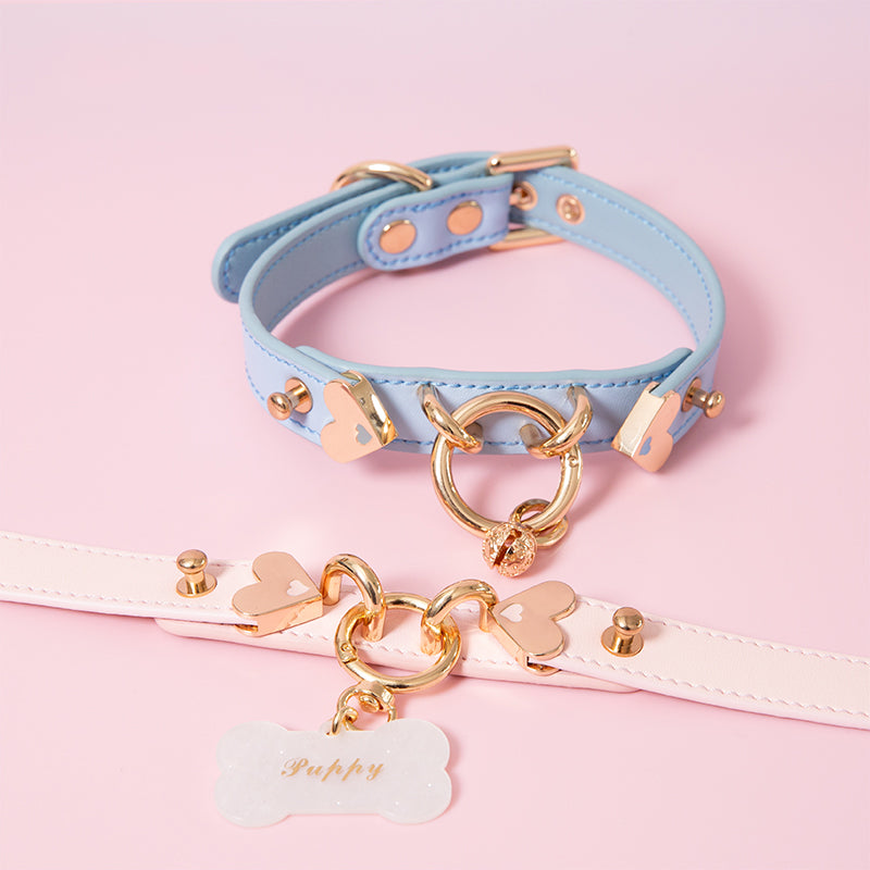 Two leather chokers in baby blue and light pink with light gold tone hardware, including a central silver O ring, heart shaped studs, detachable bell charm and detachable bone shaped pet tag, with custom engraving available for the pet tag. Displayed against a light pink background for a kawaii aesthetic.