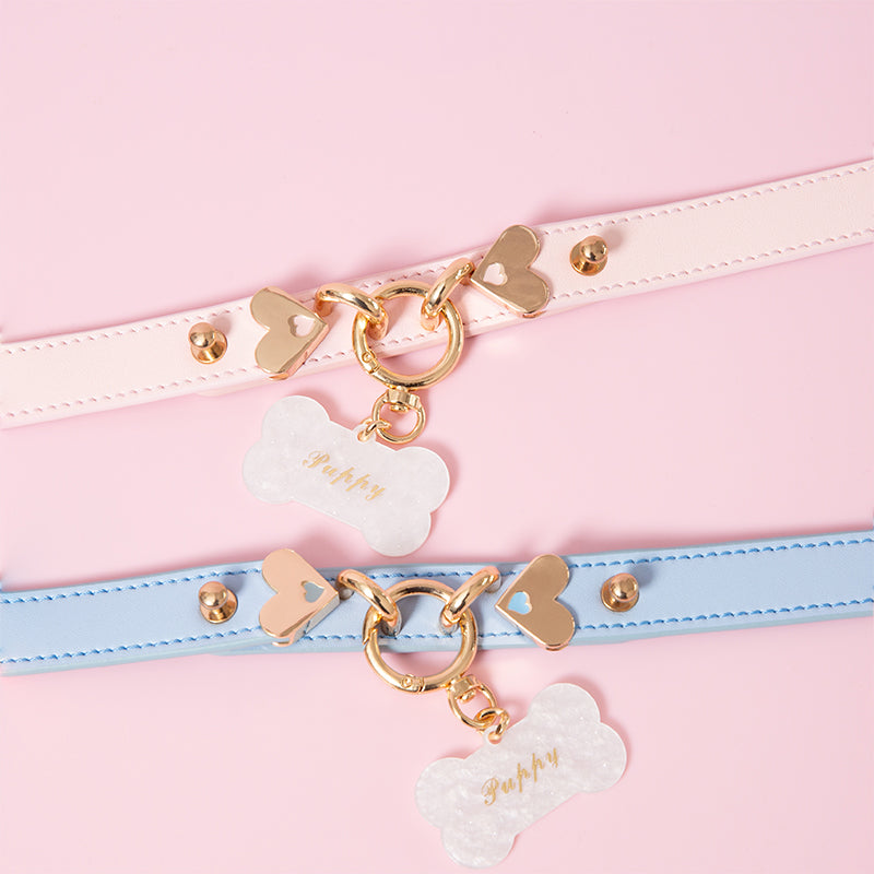Close-up of two leather chokers in baby blue and light pink with light gold tone hardware, including a central silver O ring, heart shaped studs and detachable bone shaped pet tag, with custom engraving available for the pet tag. Displayed against a light pink background for a kawaii aesthetic.