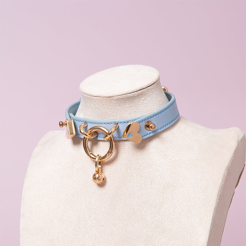 Blue leather choker featuring a light gold tone central O ring, heart shaped hardare and detachable bell charm. Displayed on a white mannequin bust against a pink background for a kawaii aesthetic.
