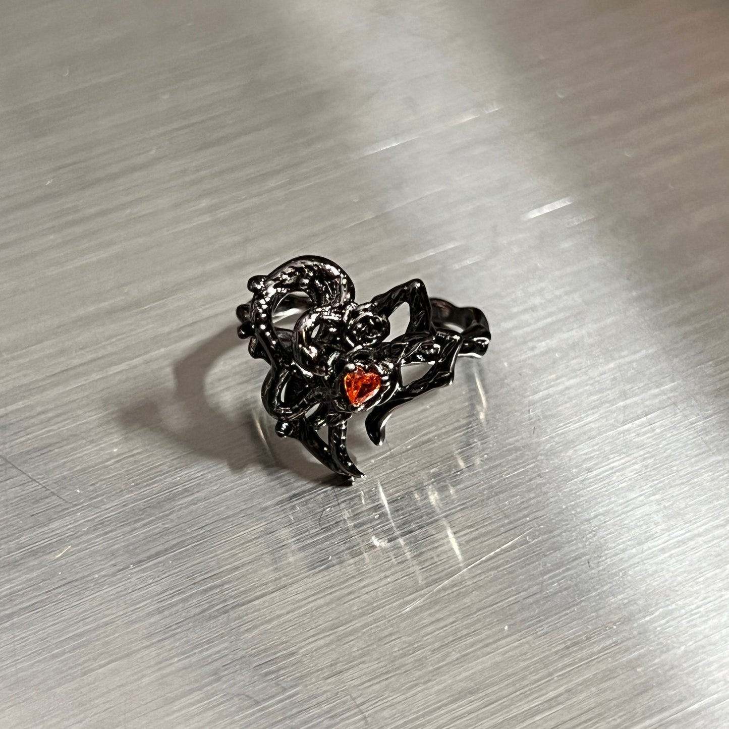 A black gothic-style ring featuring a thorny heart design with a small red heart-shaped gemstone, resting on a brushed silver metal surface.