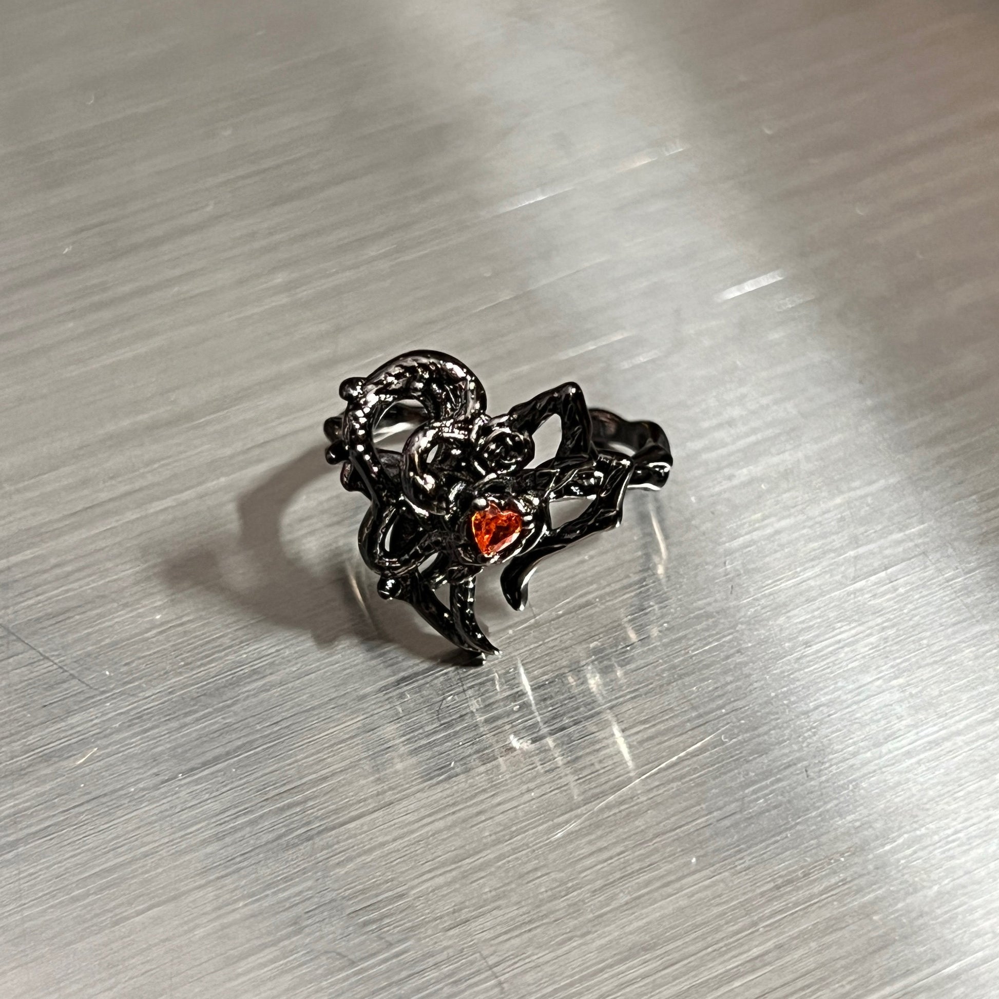 A black gothic-style ring featuring a thorny heart design with a small red heart-shaped gemstone, resting on a brushed silver metal surface.