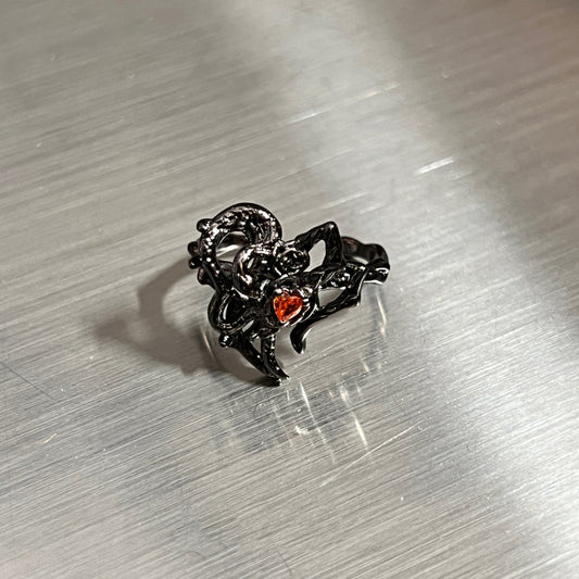 A black gothic-style ring featuring a thorny heart design with a small red heart-shaped gemstone, resting on a brushed silver metal surface.