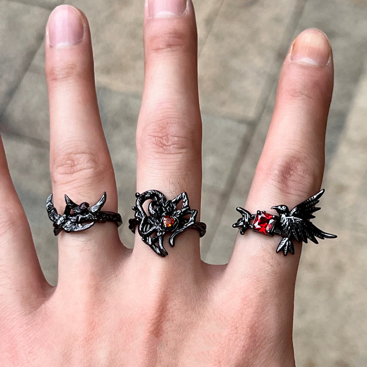 A black gothic-style ring featuring a thorny heart design with a small red heart-shaped gemstone, worn on a hand with two other matching rings.
