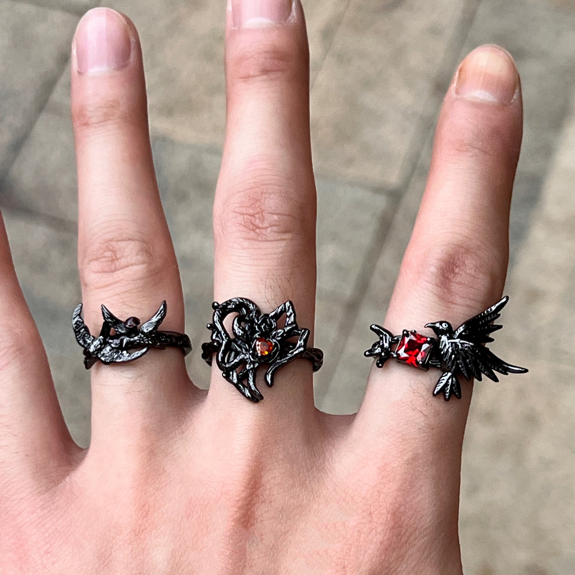 A black gothic-style ring featuring a thorny heart design with a small red heart-shaped gemstone, worn on a hand with two other matching rings.