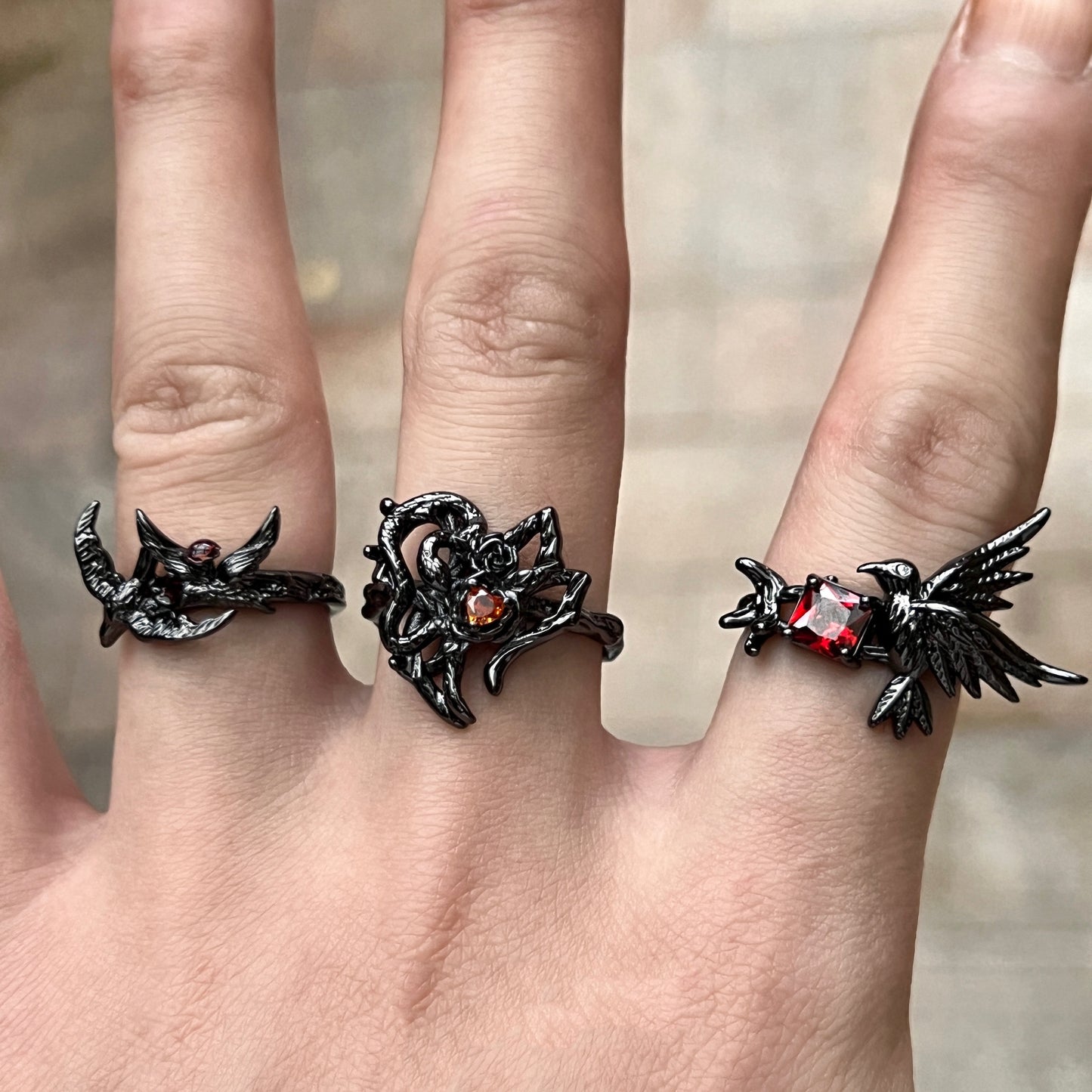 A black gothic-style ring featuring a thorny heart design with a small red heart-shaped gemstone, worn on a hand with two other matching rings.