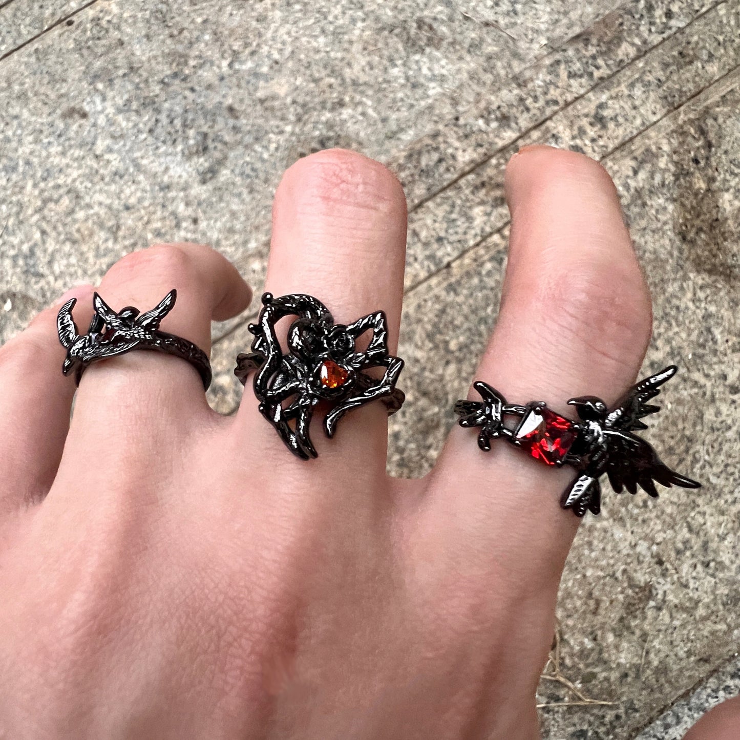 A black gothic-style ring featuring a thorny heart design with a small red heart-shaped gemstone, worn on a hand with two other matching rings.