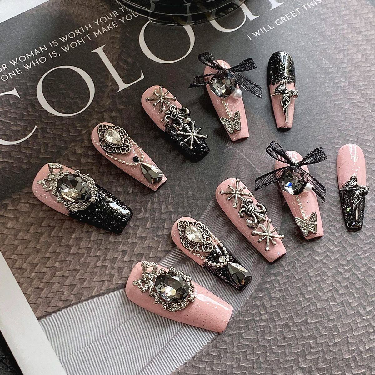 A set of coffin-shaped press-on nails displayed on a textured gray magazine page. The nails feature a pink glitter base with various embellishments including black glitter sections, black ribbon bows, silver filigree, butterfly charms, and large clear rhinestones.