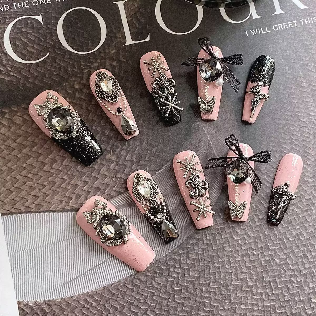 A set of decorative press-on nails displayed on a textured gray surface with a magazine page in the background. The nails are primarily light pink and black with ornate silver glitter designs, rhinestones, bows, and butterfly embellishments.