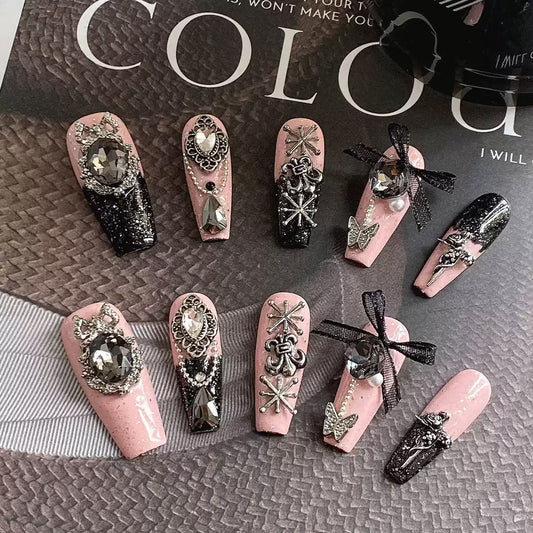 A set of decorative press-on nails displayed on a textured gray surface with a magazine page in the background. The nails are primarily light pink and black with ornate silver glitter designs, rhinestones, bows, and butterfly embellishments.