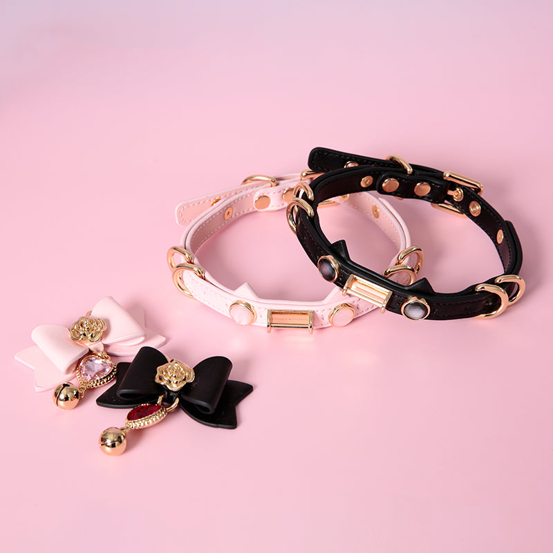 Two pink and black leather chokers featuring a cat ear shaped strap, gold tone D rings and hardware, faux mother of pearl studs and detachable leather ribbon pendants with crystal heart and gold bell charms. Displayed against a pink background for a pastel goth aesthetic.