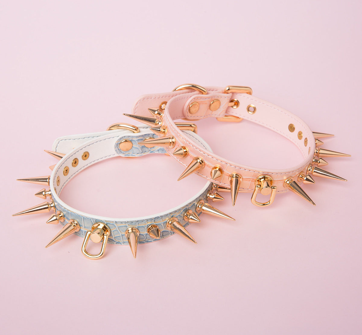 Two pink and blue faux crocodile effect leather chokers featuring gold tone alternating length spike studs and central D ring attachment. Displayed on a pink background for a Harajuku fashion aesthetic.