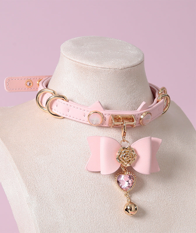 Pink leather choker featuring a cat ear shaped strap, detachable leather bow charm with a pink and gold crystal heart and bell charm. Displayed on a white mannequin bust against a pink background for a pastel goth aesthetic.