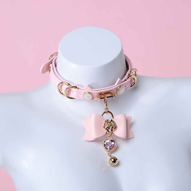 Pink leather choker featuring a cat ear shaped strap, detachable leather bow charm with a pink and gold crystal heart and bell charm. Displayed on a white mannequin bust against a pink background for a pastel goth aesthetic.