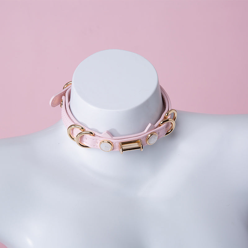 Pink leather choker featuring a cat ear shaped strap, gold tone D rings and U bar hardware and faux mother of pearl studs. Displayed on a white mannequin bust against a pink background for a Harajuku fashion aesthetic.