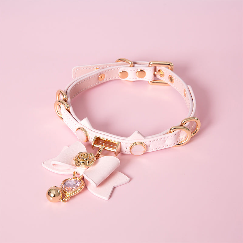 Pink leather choker featuring a cat ear shaped strap, detachable leather bow pendant with a pink and gold crystal heart and bell charm. Displayed against a pink background for a kawaii goth aesthetic.