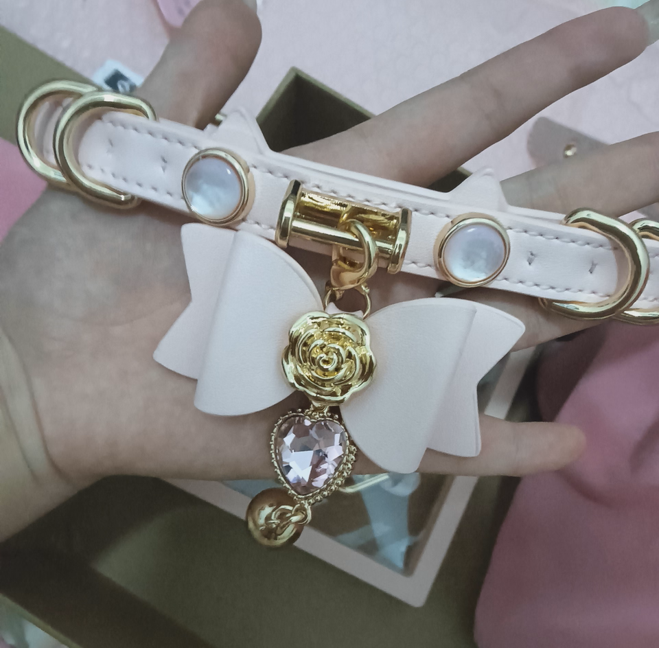 Pink leather choker featuring a cat ear shaped strap, gold tone D rings and hardware, faux mother of pearl studs and detachable pink leather bow pendant with pink and gold crystal heart and bell charms. Held by a hand against a pink background for a casual lifestyle shot.