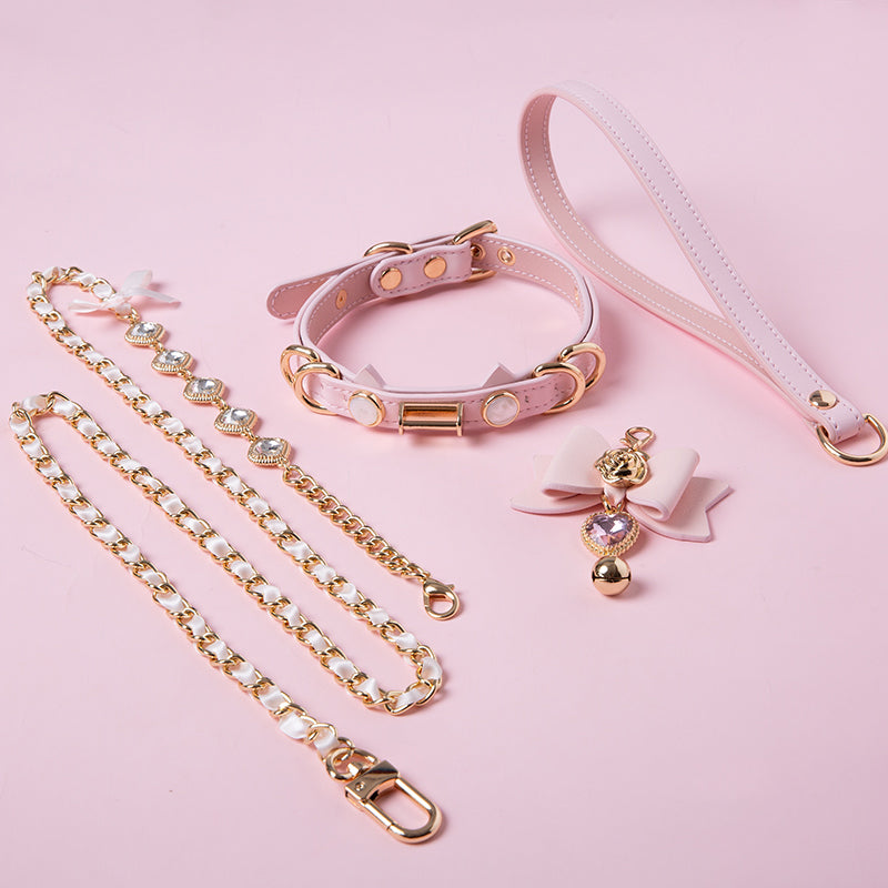 Pink leather choker and leash set featuring cat ear and ribbon shaped elements, pink and gold crystal heart and bell charm with detachable diamante, ribbon and chain leash and leather hand strap. Displayed against a pink background for a pastel goth aesthetic.