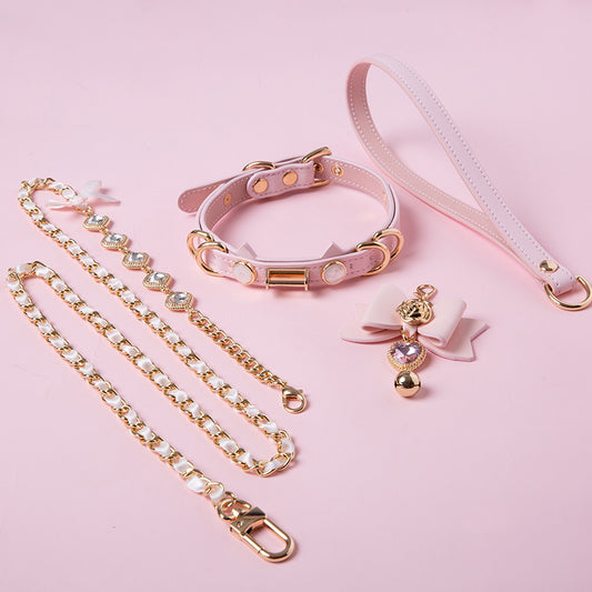 Pink leather choker and leash set featuring cat ear and ribbon shaped elements, pink and gold crystal heart and bell charm with detachable diamante, ribbon and chain leash and leather hand strap. Displayed against a pink background for a pastel goth aesthetic.