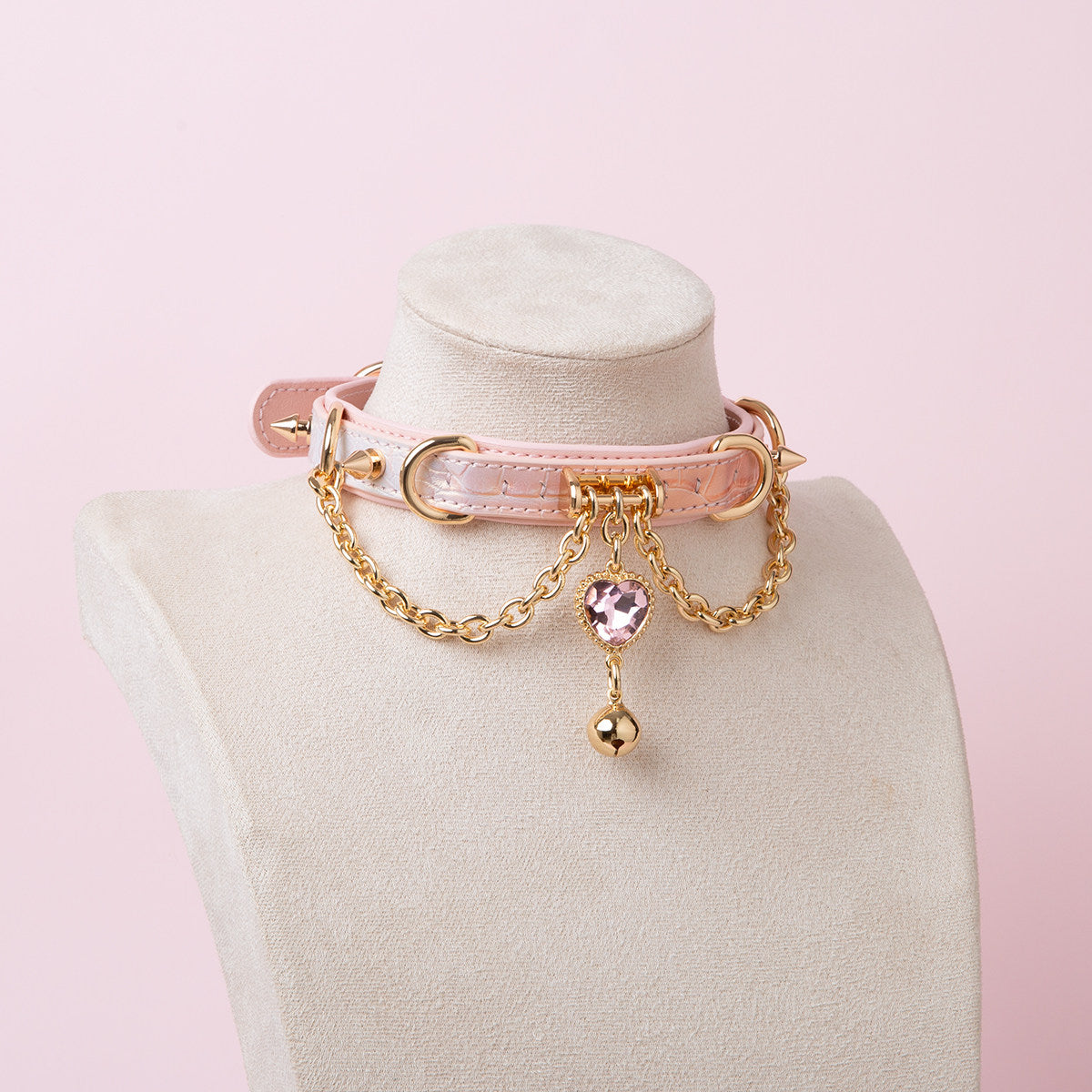Pink leather choker featuring gold tone chains, D rings, spike studs, bell and pink and gold crystal heart charm. Displayed on a white mannequin bust against a pink background for a Harajuku fashion aesthetic.